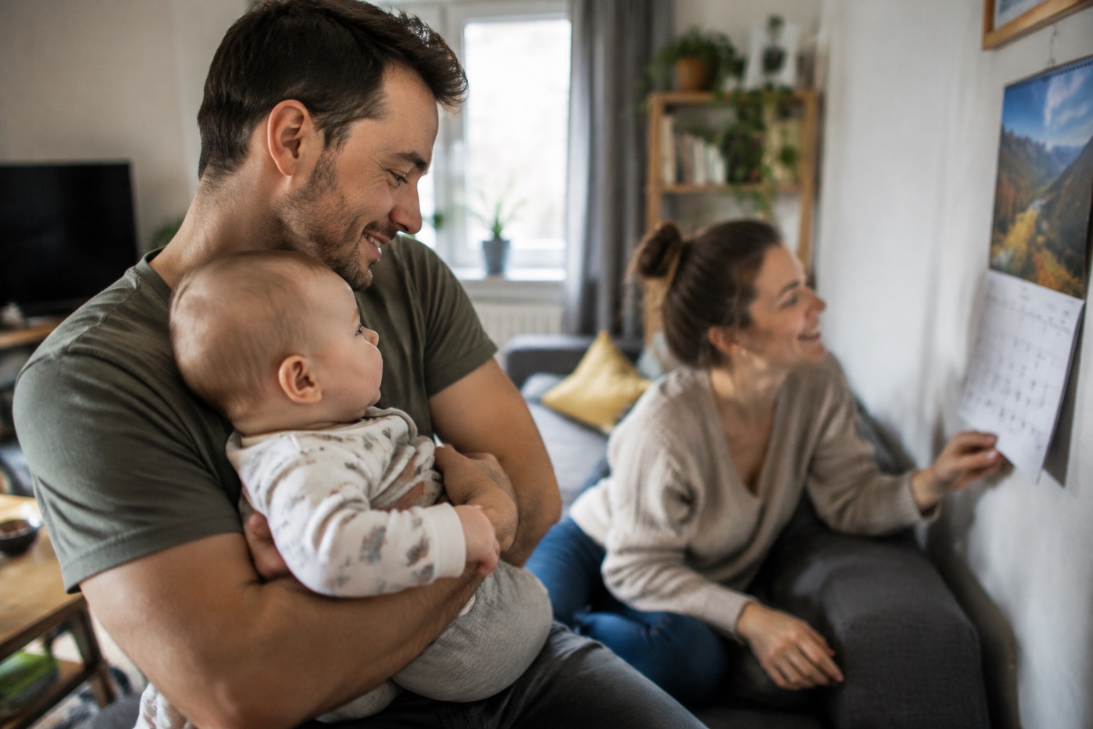 Młody tata z niemowlęciem na rękach w salonie, uśmiechnięty, obok mama przeglądająca kalendarz